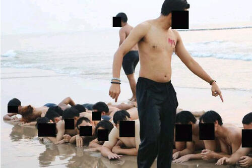 Many young male students with their faces blurred out to protect their identities lay face down on the beach as older students yell at them from above. It is not a fun scene.