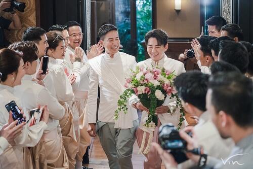 Tom Tan and Oui Srimuang, a Thai couple celebrating 20 years together, walk into the marriage hall on the day of their wedding Two men wearing traditional tops walk down the isle, surrounded by their smiling friends and family. One of them is carrying a bouquet. They are both smiling very widely.