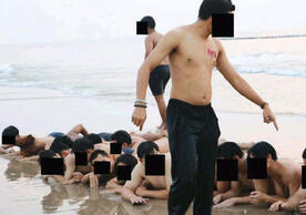 Many young male students with their faces blurred out to protect their identities lay face down on the beach as older students yell at them from above. It is not a fun scene.
