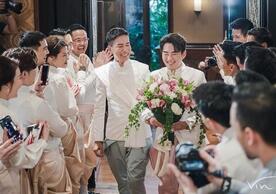 Two men wearing traditional tops walk down the isle, surrounded by their smiling friends and family. One of them is carrying a bouquet. They are both smiling very widely.